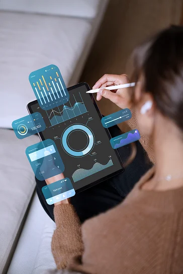 Een foto, genomen over de schouder van een vrouw die met een stylus op een tablet werkt, met een futuristisch digitaal bedieningspaneel waarop grafieken en gegevens worden weergegeven.